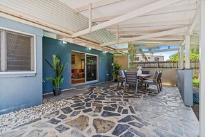 A covered patio area with stone flooring, a table with chairs, and a sliding glass door leading to an interior space. - Tropical 3-Bedroom Retreat Steps to the Beach (Holloways Beach)