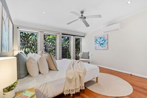 A bedroom with a bed, a ceiling fan, a window with a view of greenery, and a painting on the wall. - Tropical 3-Bedroom Retreat Steps to the Beach (Holloways Beach)