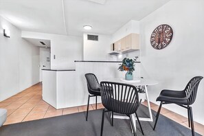 A modern kitchen with white cabinetry, a dining area with two black chairs, and a round wall clock. - Heart of the City • 1BR with Balcony & Parking (Cairns City)