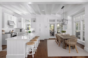 A spacious kitchen with white cabinetry, a central island with stools, a dining table with chairs, and a large window providing natural light. - Elegant Golf Course Retreat in West Wilson (Bluffton)
