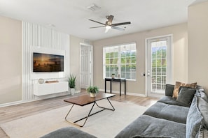 A living room with a flat-screen TV mounted on a wall, a ceiling fan, a coffee table, a sofa, and a window with blinds. - Spacious 3BR in Prime Orlando Location (Orlando)