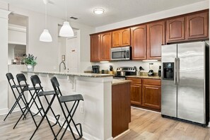 A modern kitchen with wooden cabinets, stainless steel appliances, a bar area with chairs, and pendant lighting. - Spacious 3BR in Prime Orlando Location (Orlando)