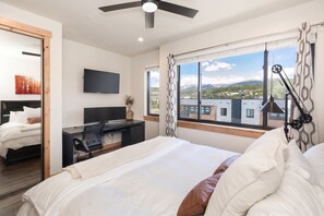 A hotel room with a bed, a desk with a chair, a flat-screen TV, and a large window with a view of buildings and mountains. - Arrow Stadthaus | Stargaze From The Private Hot Tub | Pet Friendly | Modern Downtown Townhome (Winter Park)