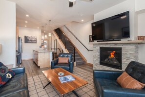 A modern living room with a fireplace, a flat-screen TV, leather sofas, and a kitchen in the background. - Arrow Stadthaus | Stargaze From The Private Hot Tub | Pet Friendly | Modern Downtown Townhome (Winter Park)