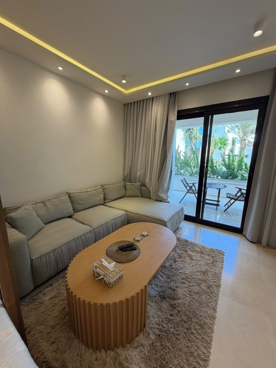 Una sala de estar moderna con un sofá modular, una mesa redonda y un amplio ventanal corredizo que da a un espacio exterior.