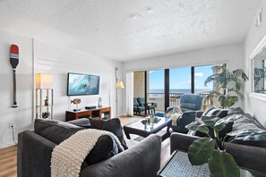 A modern living room with a large TV, comfortable seating, a balcony with a view, and a decorative wall. - Seventh Heaven: 7th-Floor Ocean View Condo with Beach Access & Heated Pool in Cocoa Beach (Cocoa Beach)