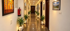 A hotel hallway with framed artwork on the walls, potted plants along the floor, and a fire extinguisher mounted on the wall. - Hotel silver stay delhi  (New Delhi)