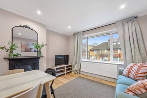 A modern living room with a flat-screen TV, a dining table, a sofa, and a large window with curtains. - Lovely Ealing 3-bed in Greater London (London)
