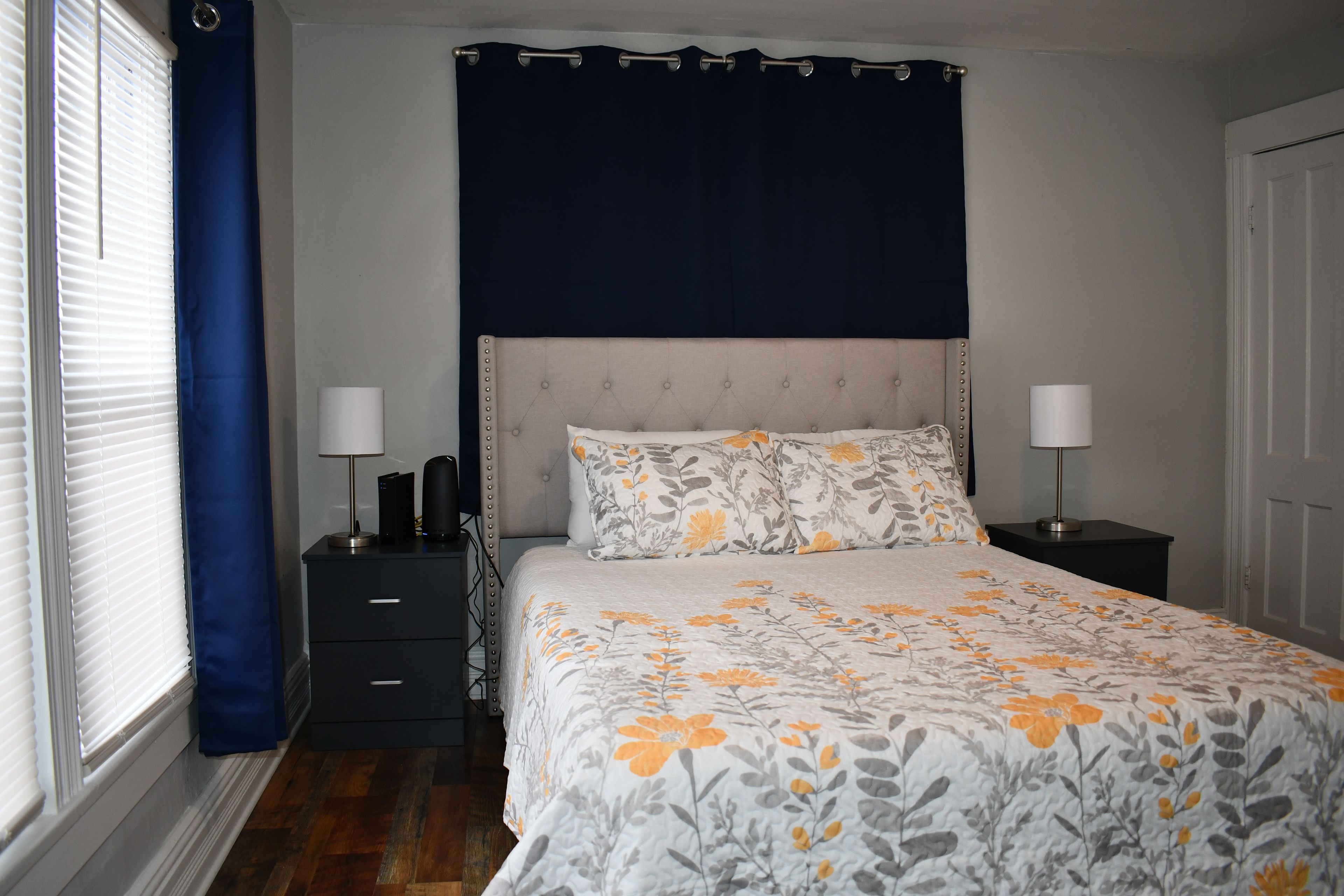 A bedroom with a bed, two bedside tables, a nightstand, a window with blinds, and a curtain.