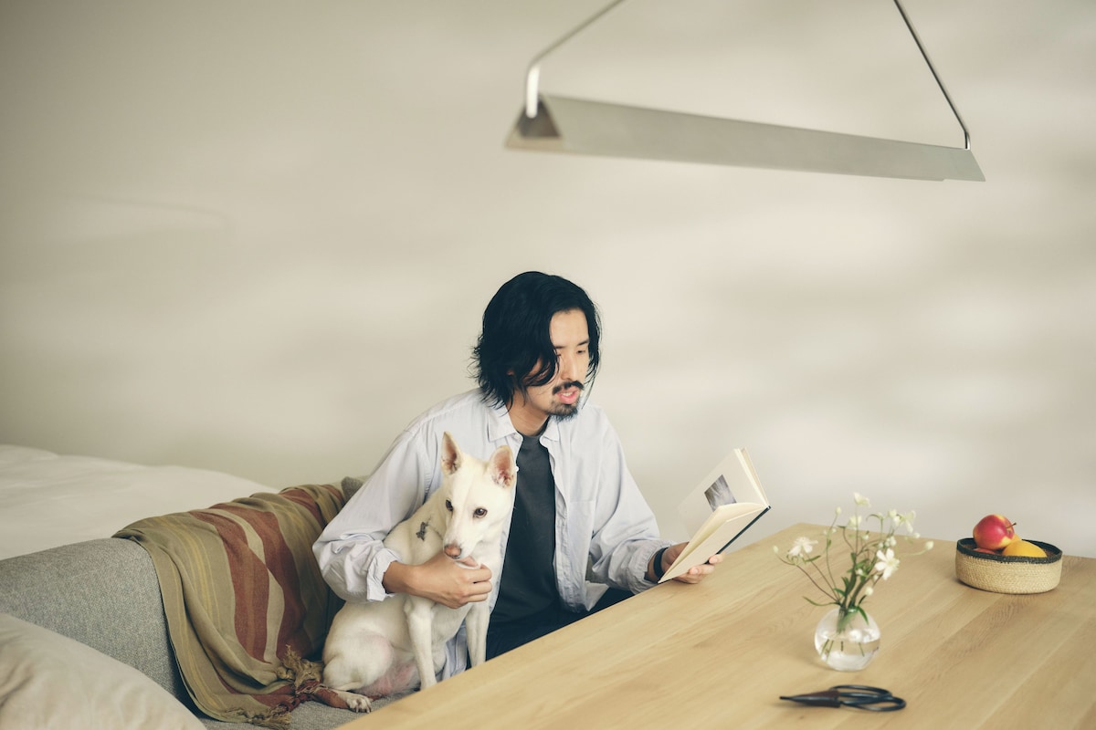 A person reading a book at a wooden table with a dog, a vase with flowers, and a basket with fruit.