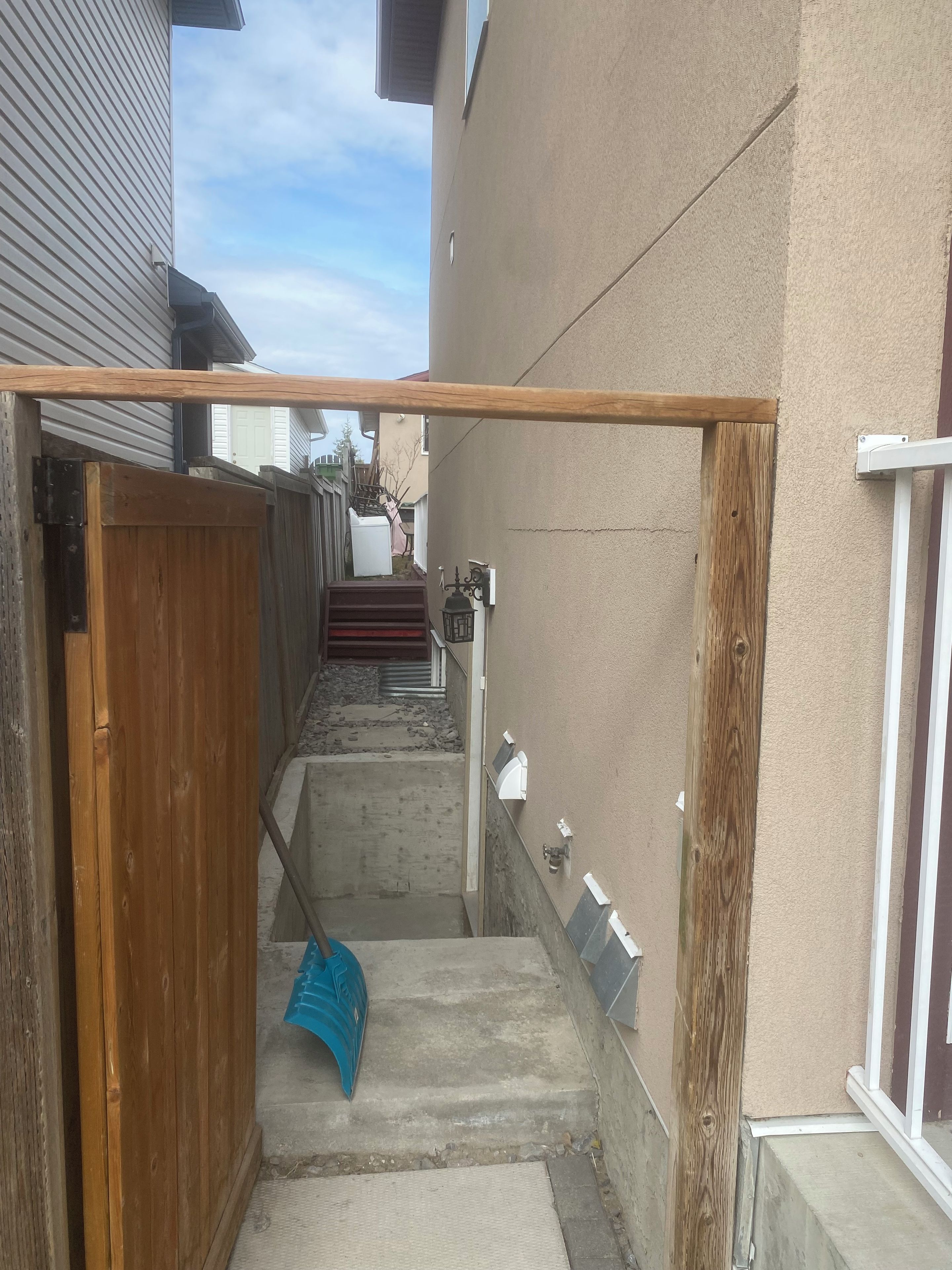 Un étroit sentier extérieur mène à une porte en bois, avec une marche en béton et une main courante en métal.
