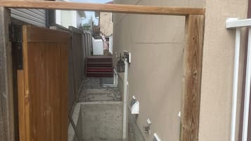 Un étroit sentier extérieur mène à une porte en bois, avec une marche en béton et une main courante en métal.