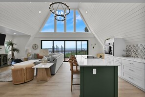Amplia sala de estar con techo alto, un ventanal grande con vista a un cuerpo de agua y una cocina moderna con armarios blancos.