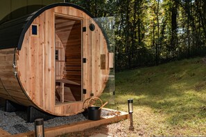 Estructura de sauna de madera con puerta abierta, un pequeño balde negro y un banco de madera en el exterior.