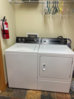 A laundry area with two white front-loading washing machines, a fire extinguisher, and a metal drying rack with colorful hangers. - Superb waterfront 3-bedroom 2 1/2 bath townhome in charming Sitka by the Sea, (Sitka)