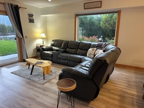 A living room with a leather sofa, a wooden coffee table, and a view of greenery outside. - Superb waterfront 3-bedroom 2 1/2 bath townhome in charming Sitka by the Sea, (Sitka)