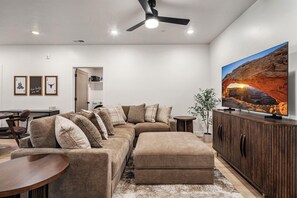 A living room with a sectional sofa, ottoman, a flat-screen TV, a ceiling fan, and a large window with a desert landscape view. - Terra P-11, Private Spa, Community Amenities w/ Water Slide! (Hurricane)