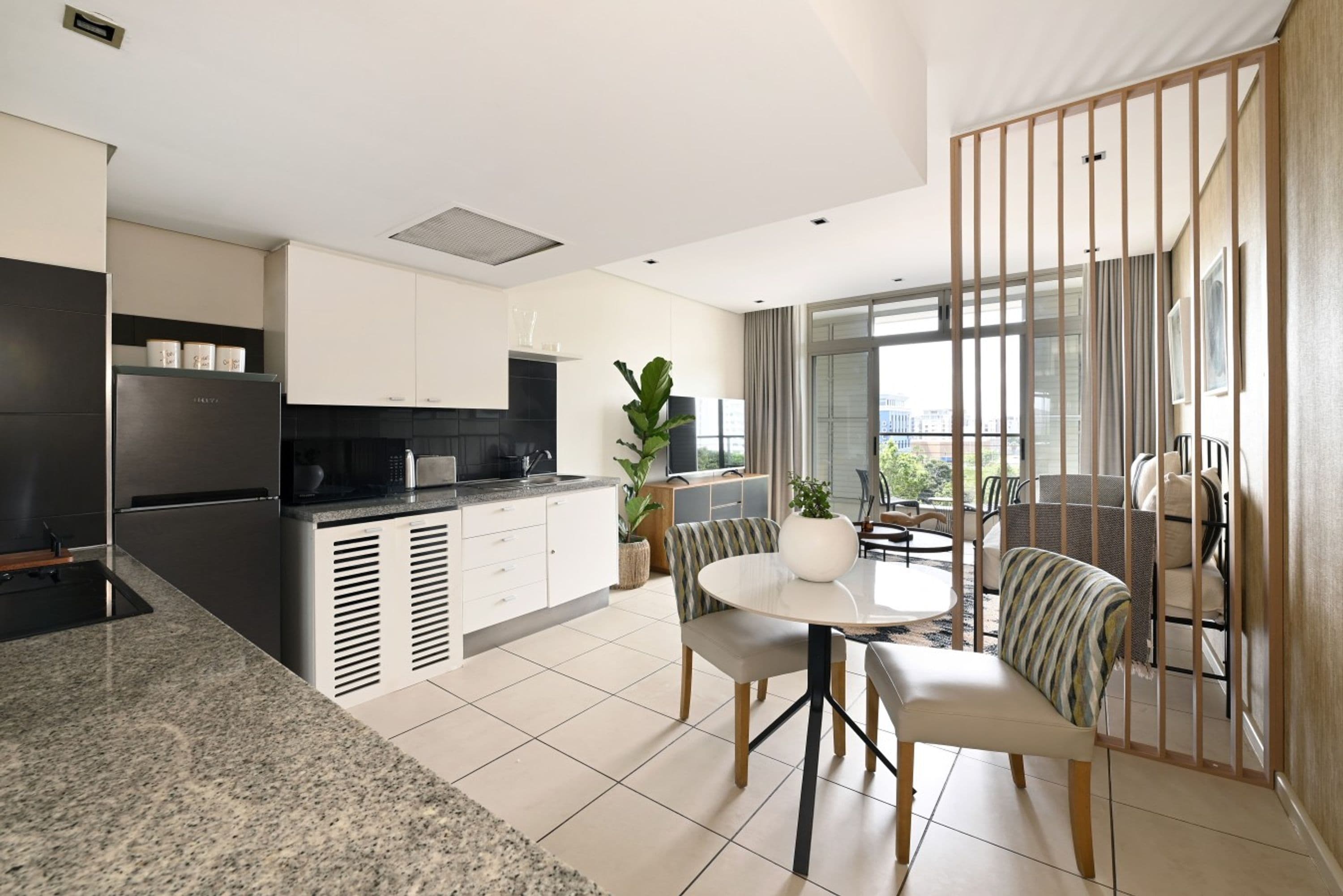 A modern kitchen with a built-in oven, a dining area with a round table and chairs, and a view of the city through large windows.