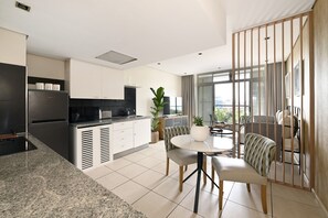 A modern kitchen with a built-in oven, a dining area with a round table and chairs, and a view of the city through large windows. - Secure One Bed Apartment Modern and Spacious (Cape Town)
