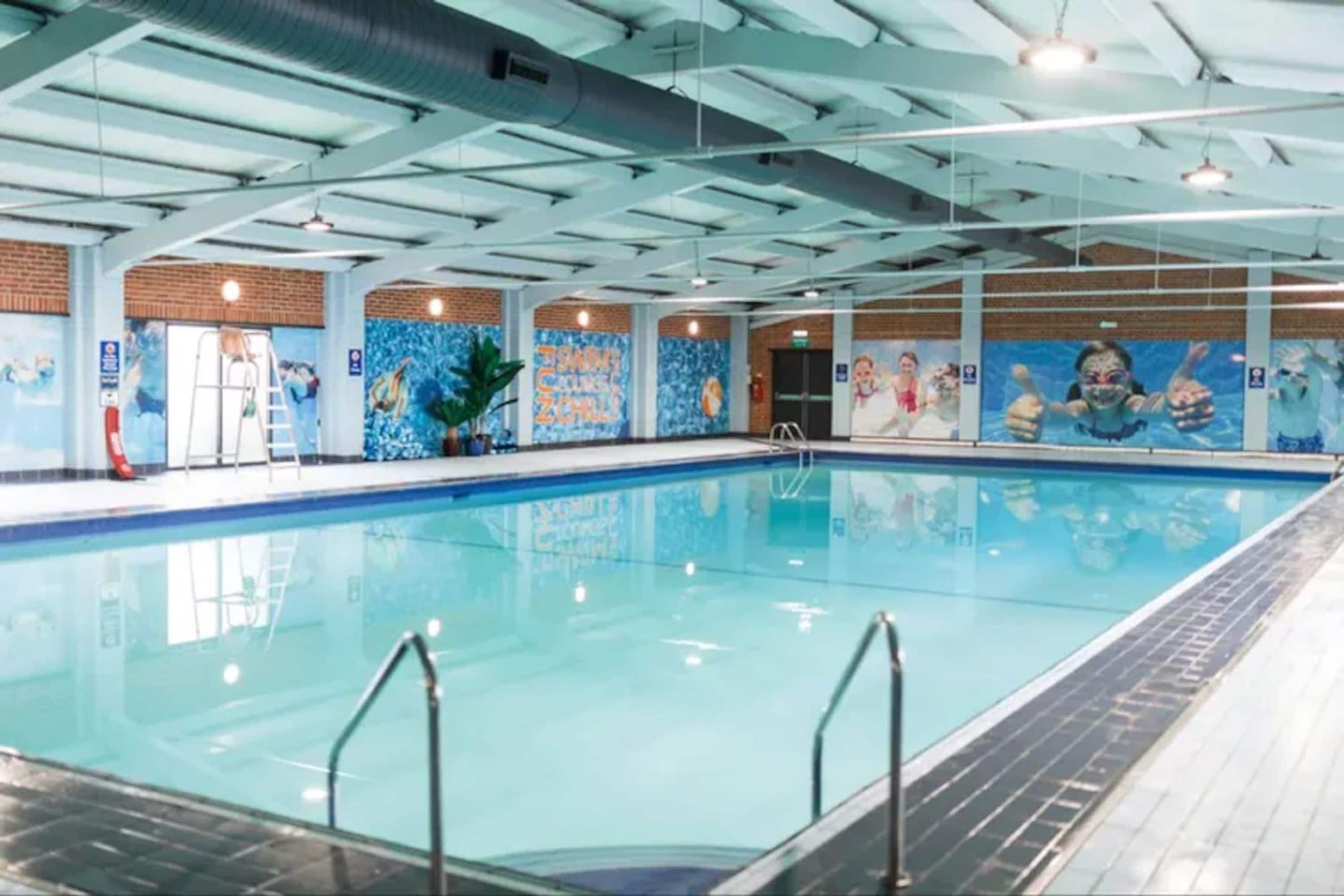 Piscina interna com água cristalina, cercada por paredes com murais e teto alto com vigas aparentes.