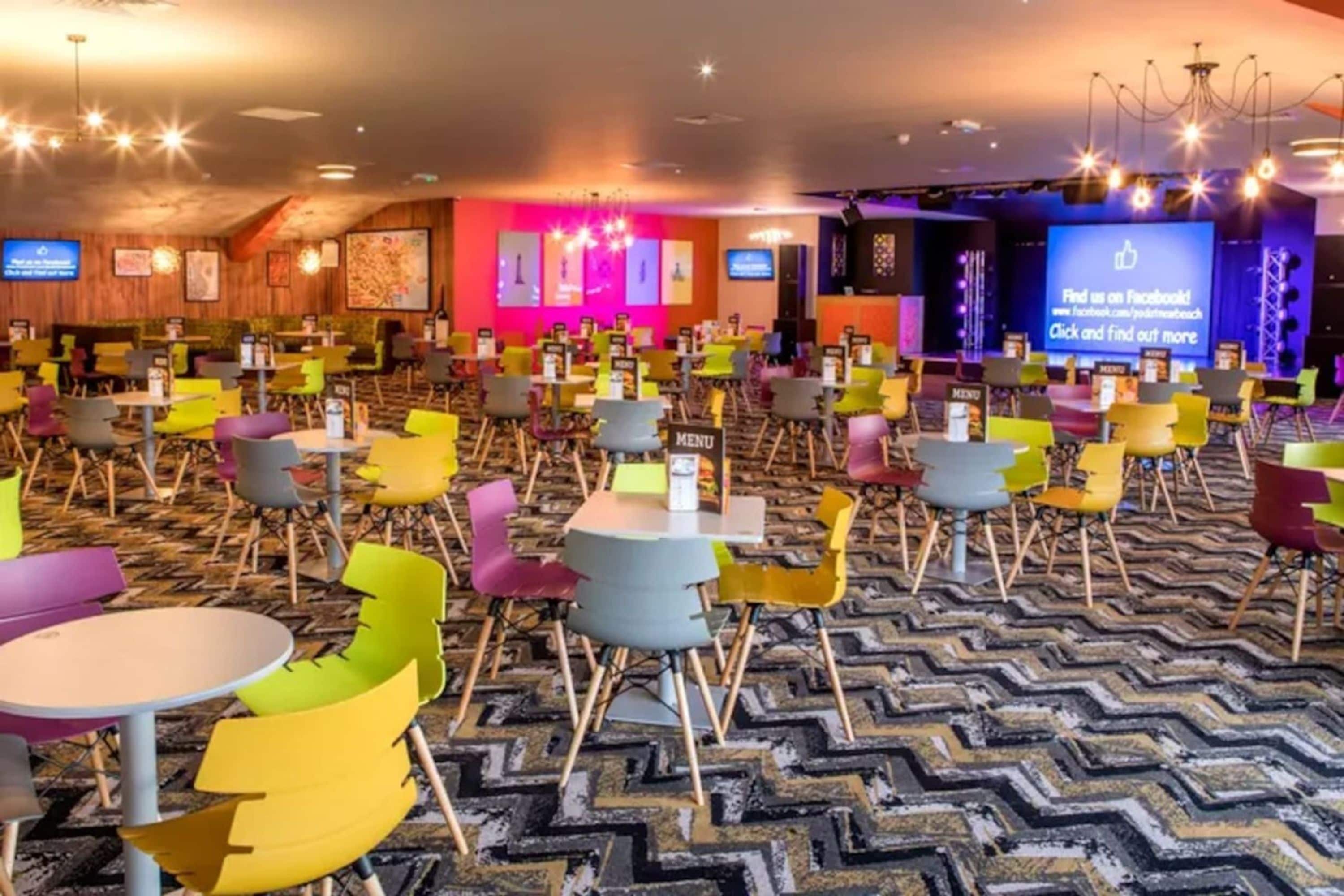 A conference room with multiple tables and chairs, a stage with a screen, and a patterned carpet.