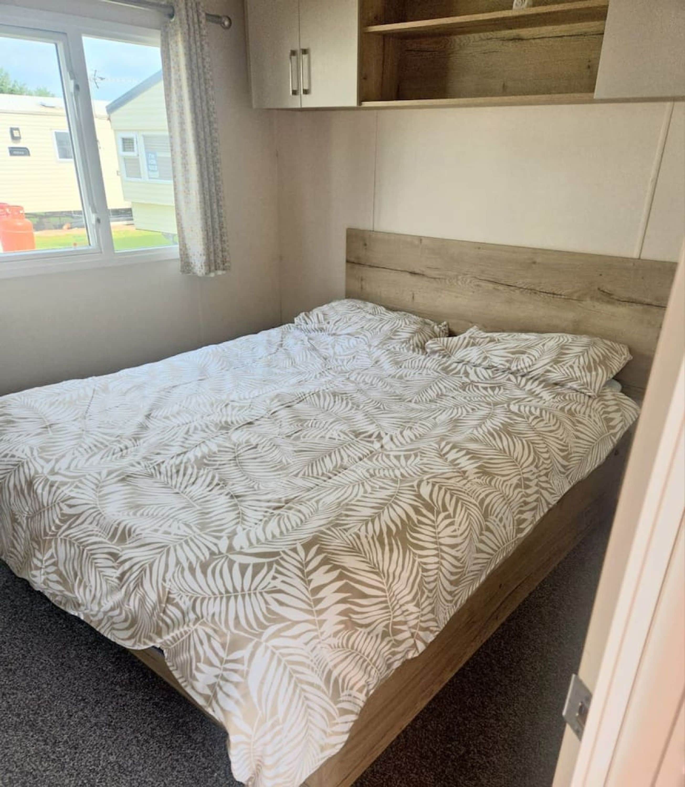 A single bed with a patterned bedspread in a room with a window, a wooden headboard, and a small shelf above the bed.
