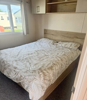 A single bed with a patterned bedspread in a room with a window, a wooden headboard, and a small shelf above the bed. - Located at New Beach Holiday Park in Dymchurch (Romney Marsh)