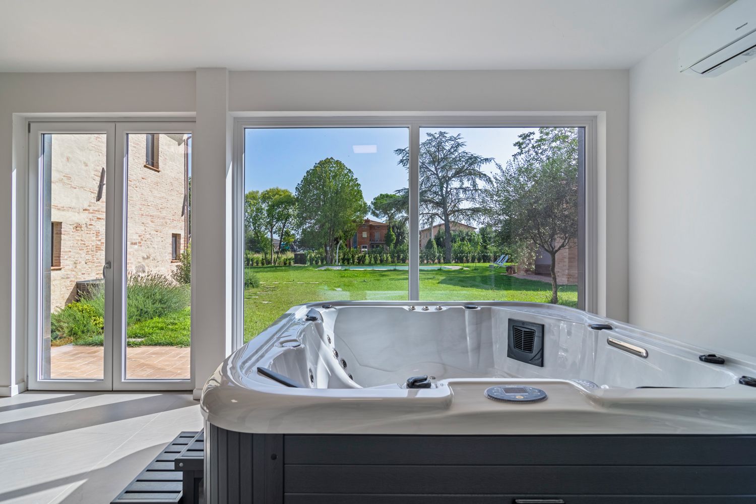 Un baño moderno con jacuzzi, ventanas amplias y vistas a un jardín y edificios.