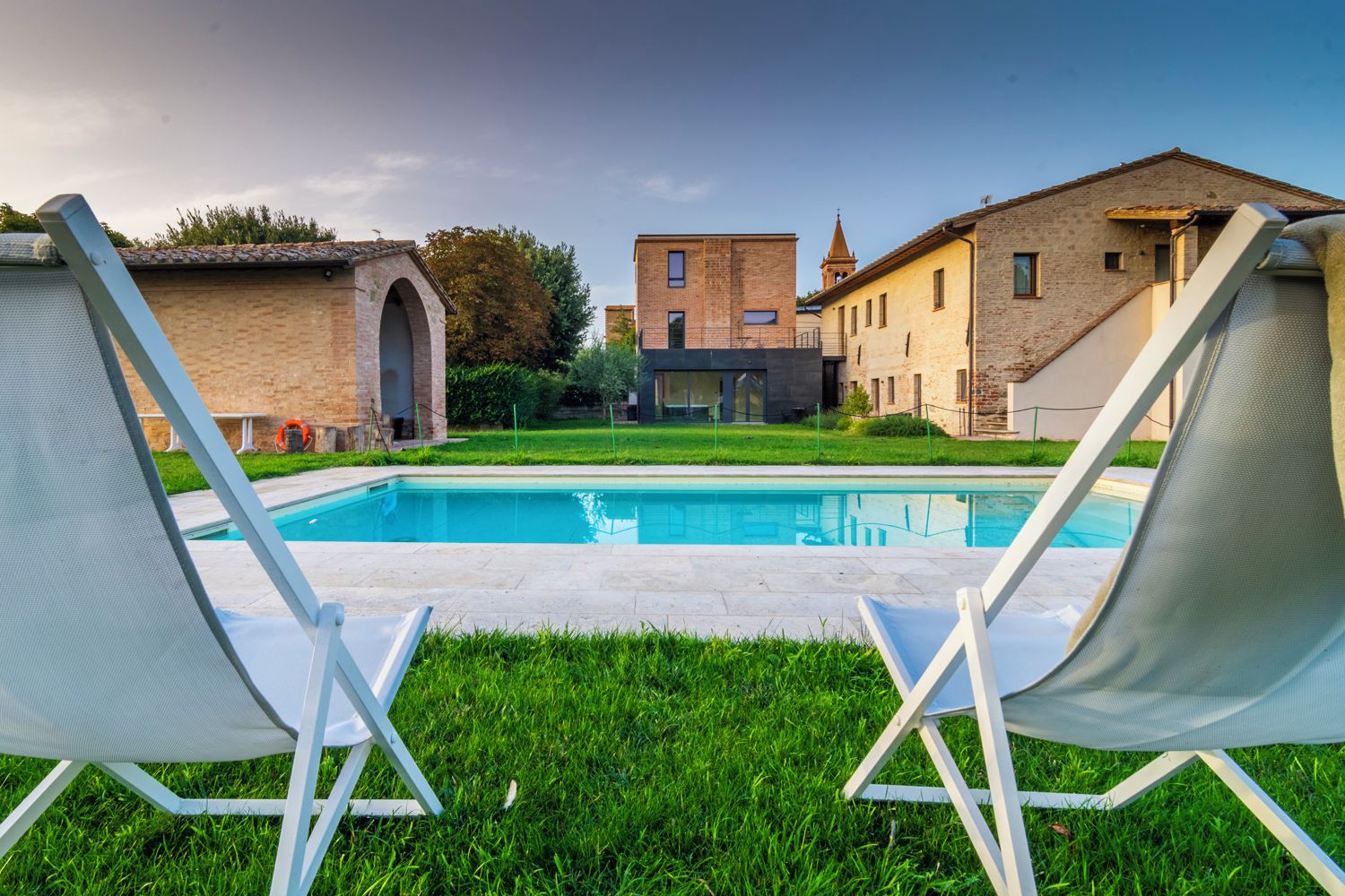 A swimming pool with two lounge chairs facing it, surrounded by a grassy area and buildings with a mix of modern and traditional architecture.