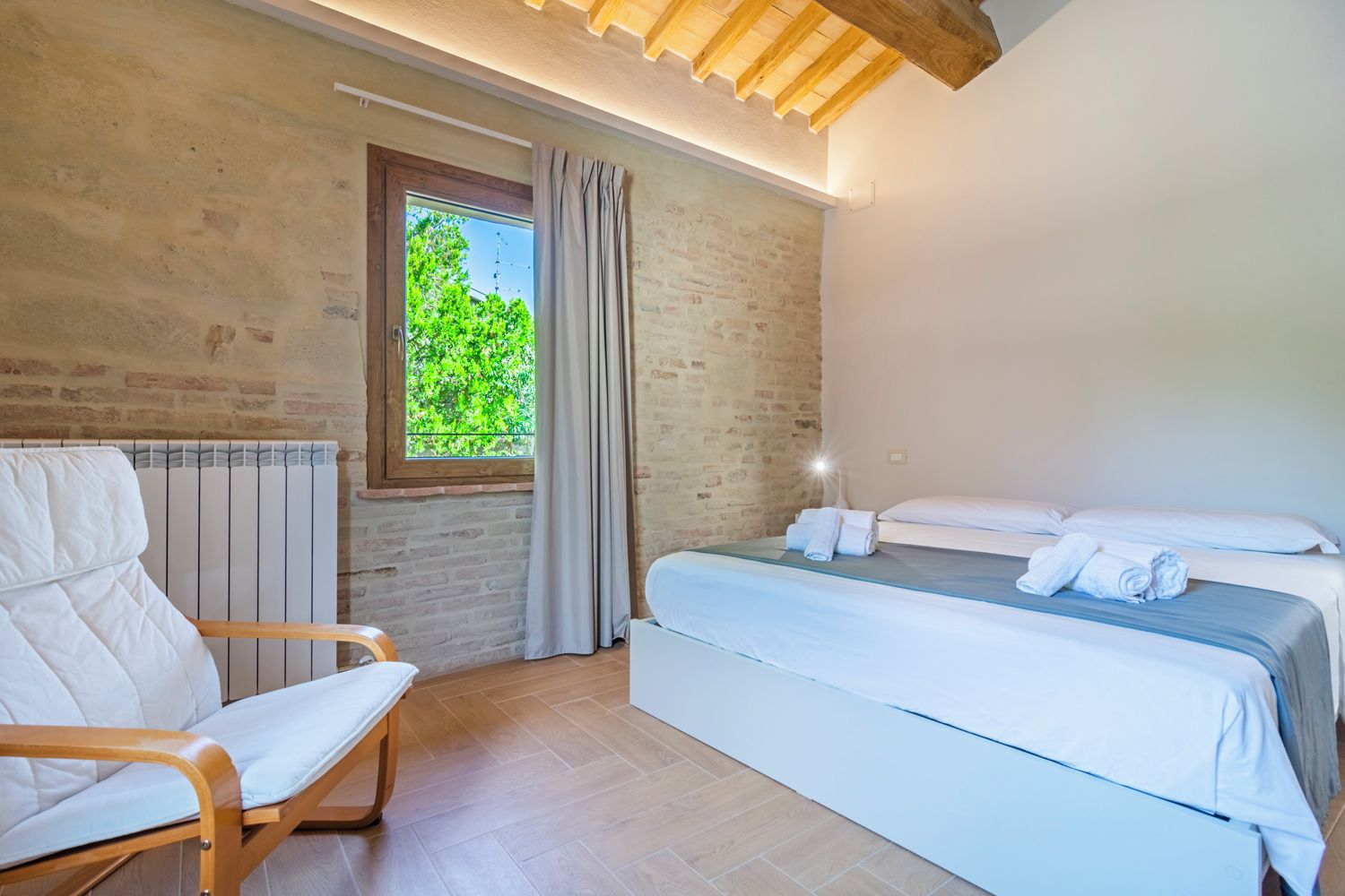 A bedroom with a bed, a chair, a window with curtains, and a brick wall.