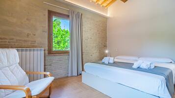 A bedroom with a bed, a chair, a window with curtains, and a brick wall.