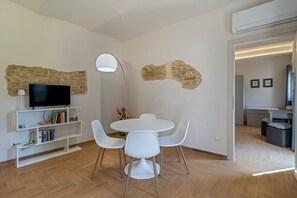 Ein modernes Wohnzimmer mit Fernseher, Bücherregalen, einem Esstisch mit Stühlen und freiliegenden Backsteinwänden.