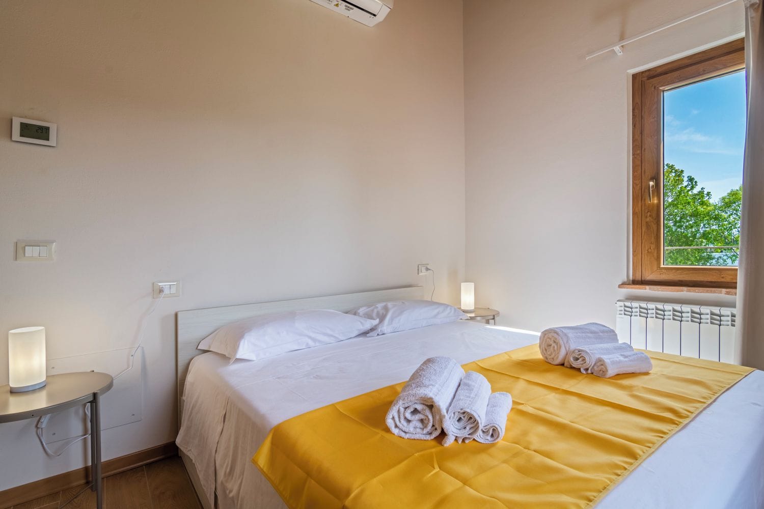 Ein Hotelzimmer mit Bett, Nachttisch, Lampe und Fenster mit Blick auf Bäume.