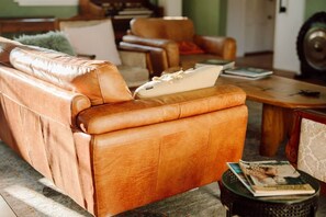 A brown leather sofa with a cushion, a small round table with a magazine, and a patterned chair in a living room.