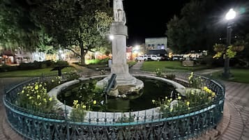 A nighttime view of a park with a statue in the center, surrounded by a circular water feature and a decorative fence.