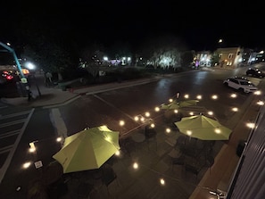 An outdoor seating area with umbrellas and tables, a street with parked cars, and buildings in the background.