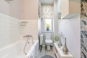 A bathroom with a bathtub, toilet, and a window with a plant. - Bogedaina Central Apartment (Chorzów)