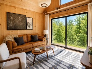 A room with a wooden interior, a leather sofa, a coffee table, and a large window with a view of greenery. - Modern Creekside Cabin w/ Hot Tub — Near Smoky's (Cosby)