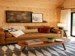 A cozy living room with a brown leather sofa, a coffee table with books and a plant, and a large framed picture on the wall. - Modern Creekside Cabin w/ Hot Tub — Near Smoky's (Cosby)