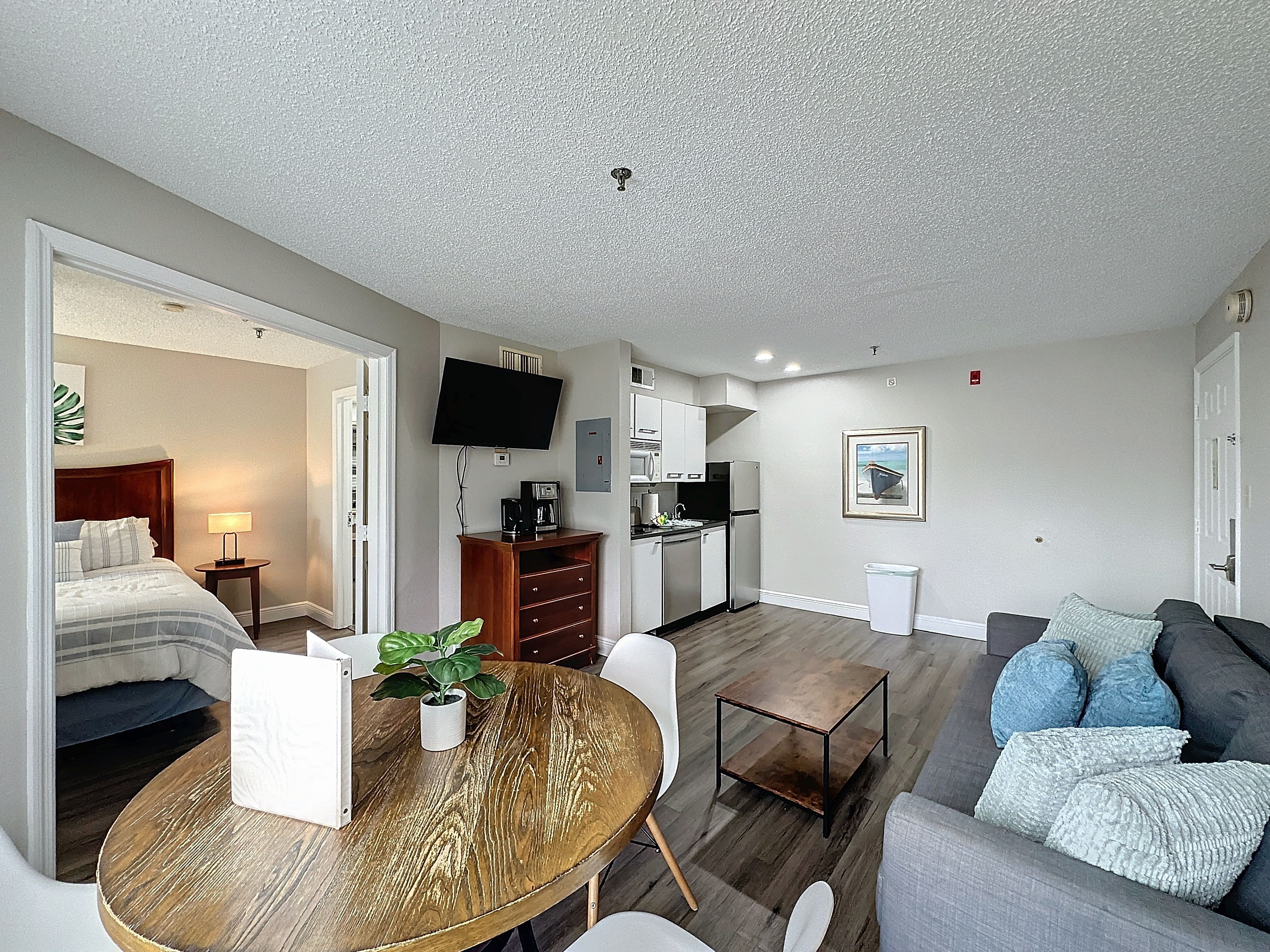 Una habitación de hotel moderna con cama, una mesa de comedor con planta, un área de estar con sofá, una cocina equipada y un televisor en la pared.
