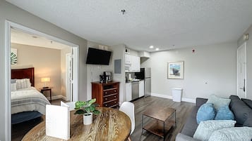 Una habitación de hotel moderna con cama, una mesa de comedor con planta, un área de estar con sofá, una cocina equipada y un televisor en la pared.