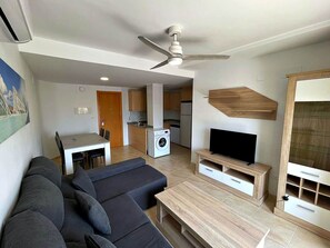 Ein modernes Wohnzimmer mit einer Couch, einem Fernseher auf einem Holzgestell, einem Deckenventilator und einer Küche im Hintergrund.