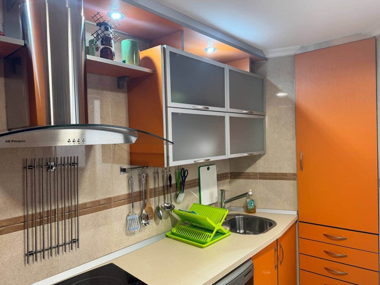 Una cocina con campana de acero inoxidable, un lavabo con un escurreplatos verde y armarios naranjas.