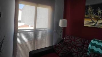 Una habitación con puerta de vidrio corredera que da a un balcón, un sofá estampado, una lámpara de pie y un cuadro en la pared.