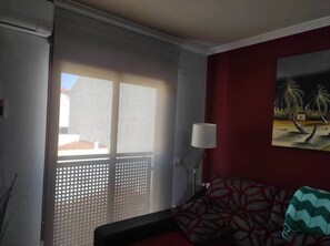 Een kamer met een schuifdeur van glas naar een balkon, een gestreept sofa, een vloerlamp en een schilderij aan de muur.