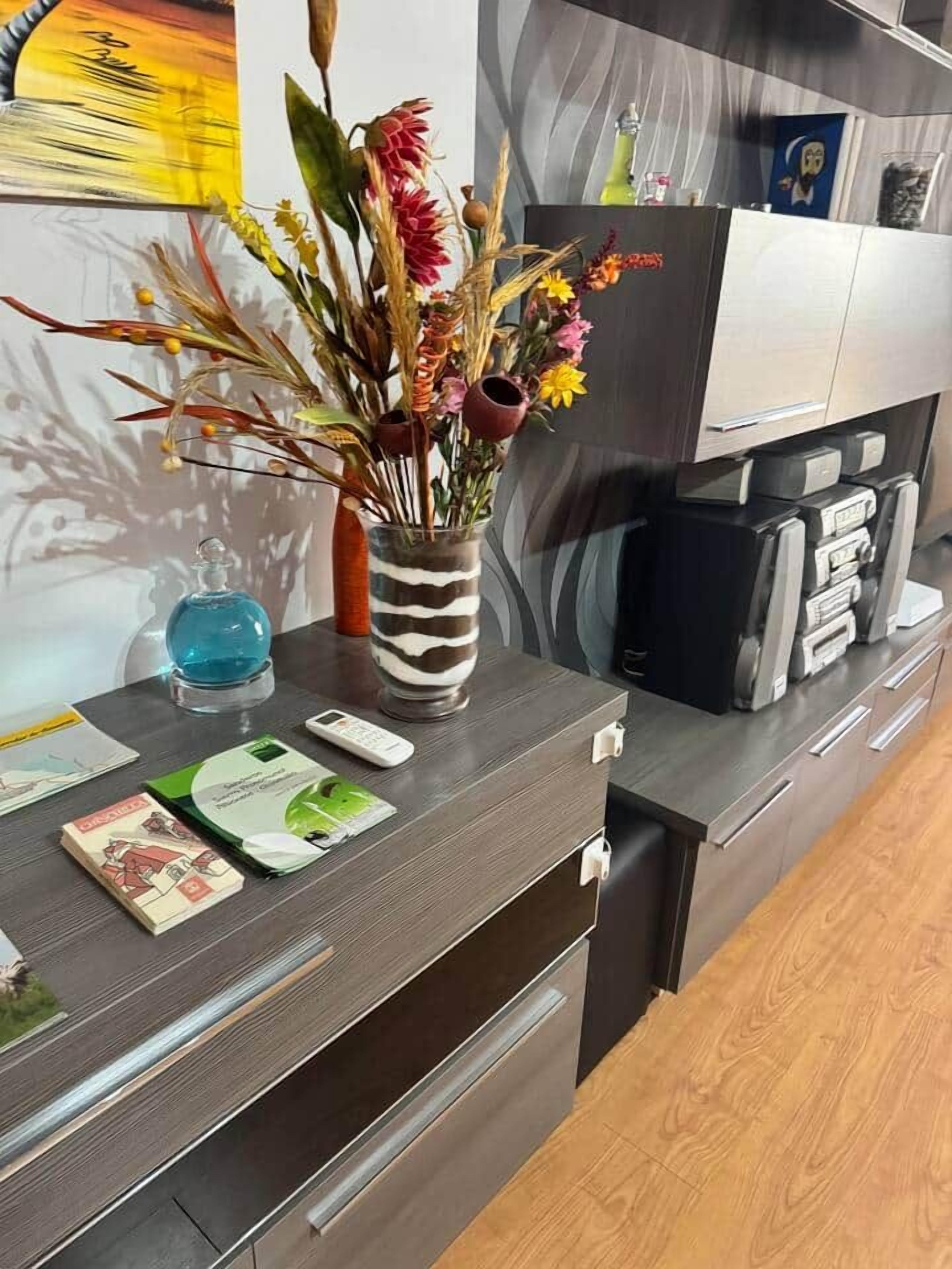 Une cuisine moderne avec un plan de travail foncé, un vase rayé contenant des fleurs séchées, une bouteille d’eau bleue et une machine à café.