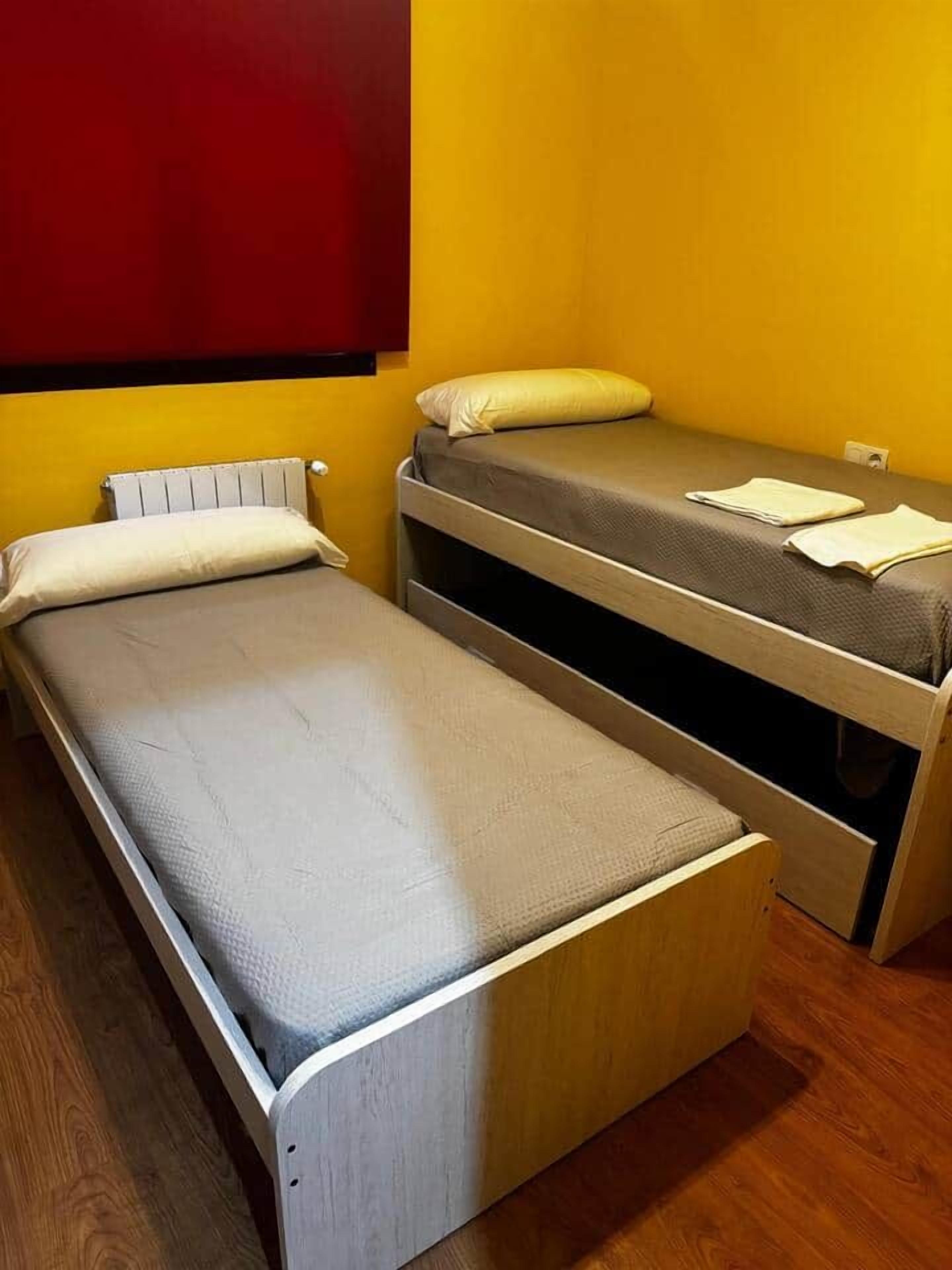A room with two beds, one with a headboard and one without, against a yellow wall.