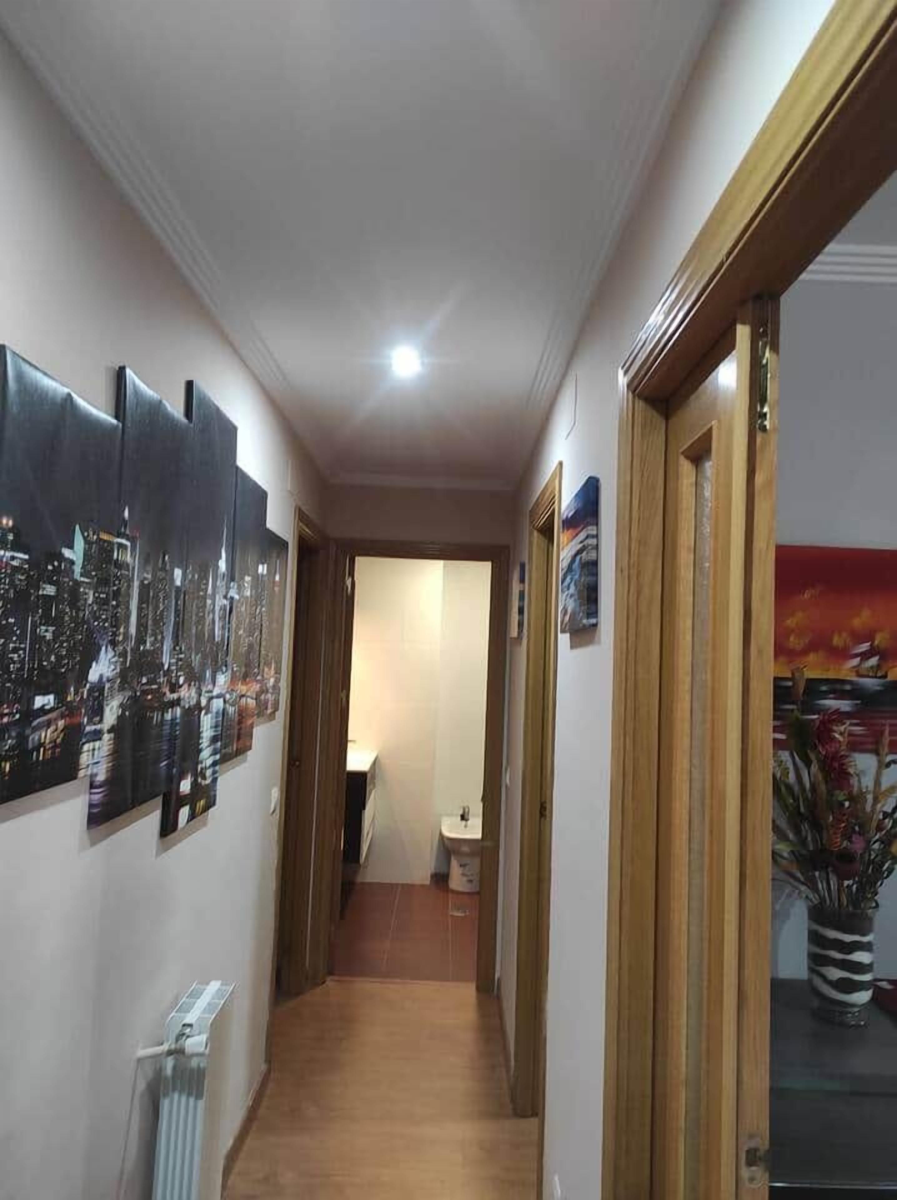 A narrow hallway with a radiator on the left, a wooden door frame on the right, and a door leading to a room with a window.