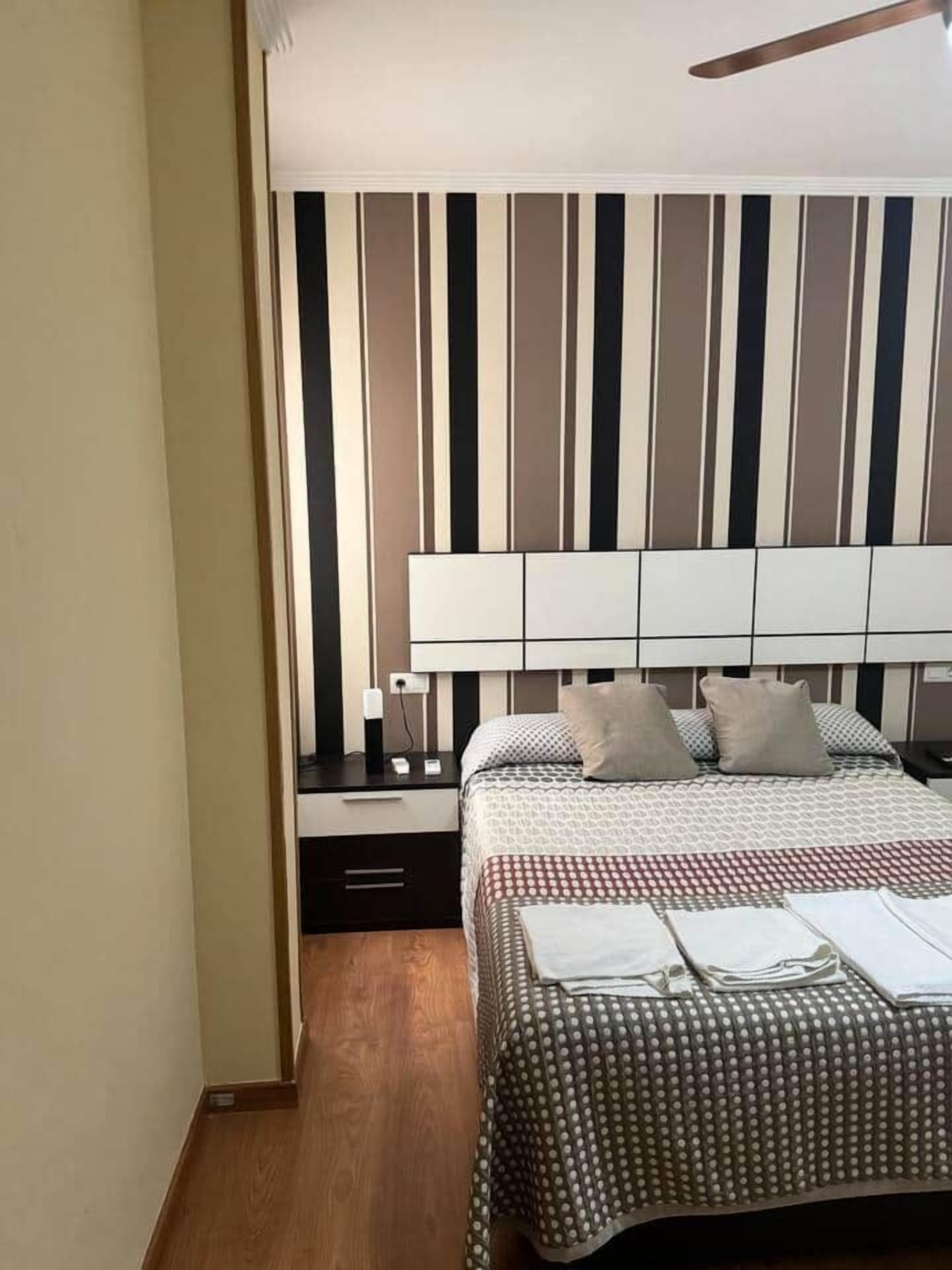 Un dormitorio con pared a rayas, cama con colcha estampada, mesita de noche con lámpara y un mueble auxiliar con teléfono.