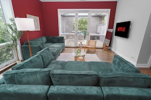 A living room with a green sofa, a TV, and a balcony with a view. - Little Italy 4BR Retreat w/ Optional Parking (Chicago)